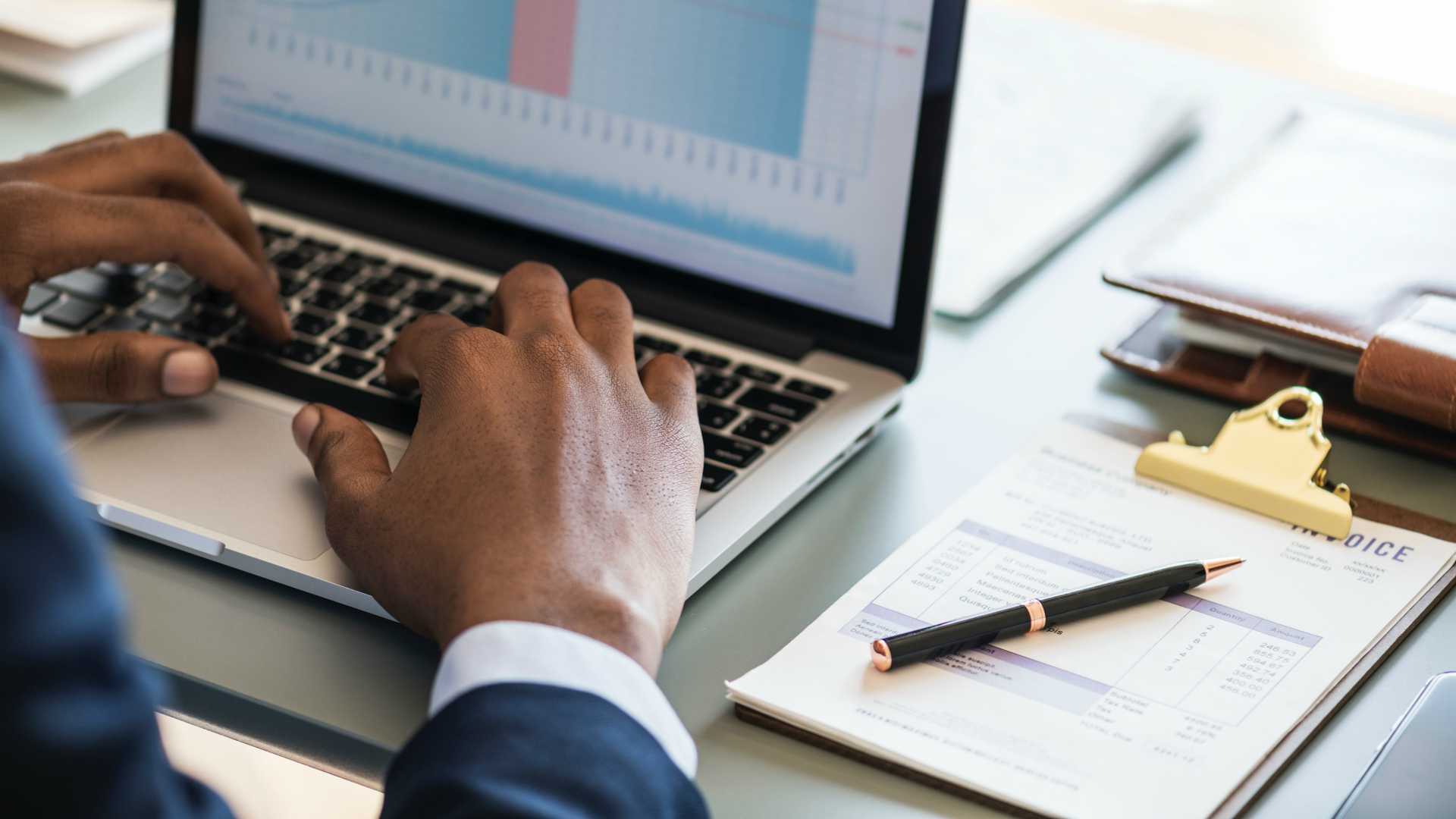 man typing on a spreadsheet with an invoice on his desk