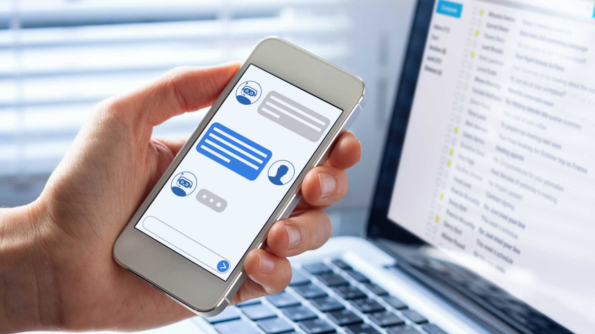person holding a smartphone with a chatbot conversation displayed on the screen; sitting in front of a laptop showing an inbox