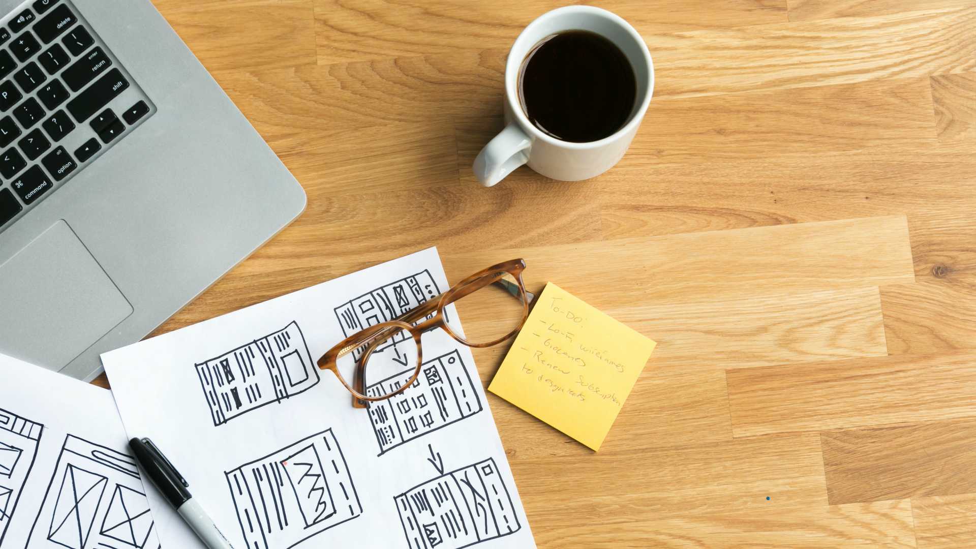 desk with handwritten module designs, post-it notes, glasses, and a cup of coffee; part of a laptop is visible in the corner