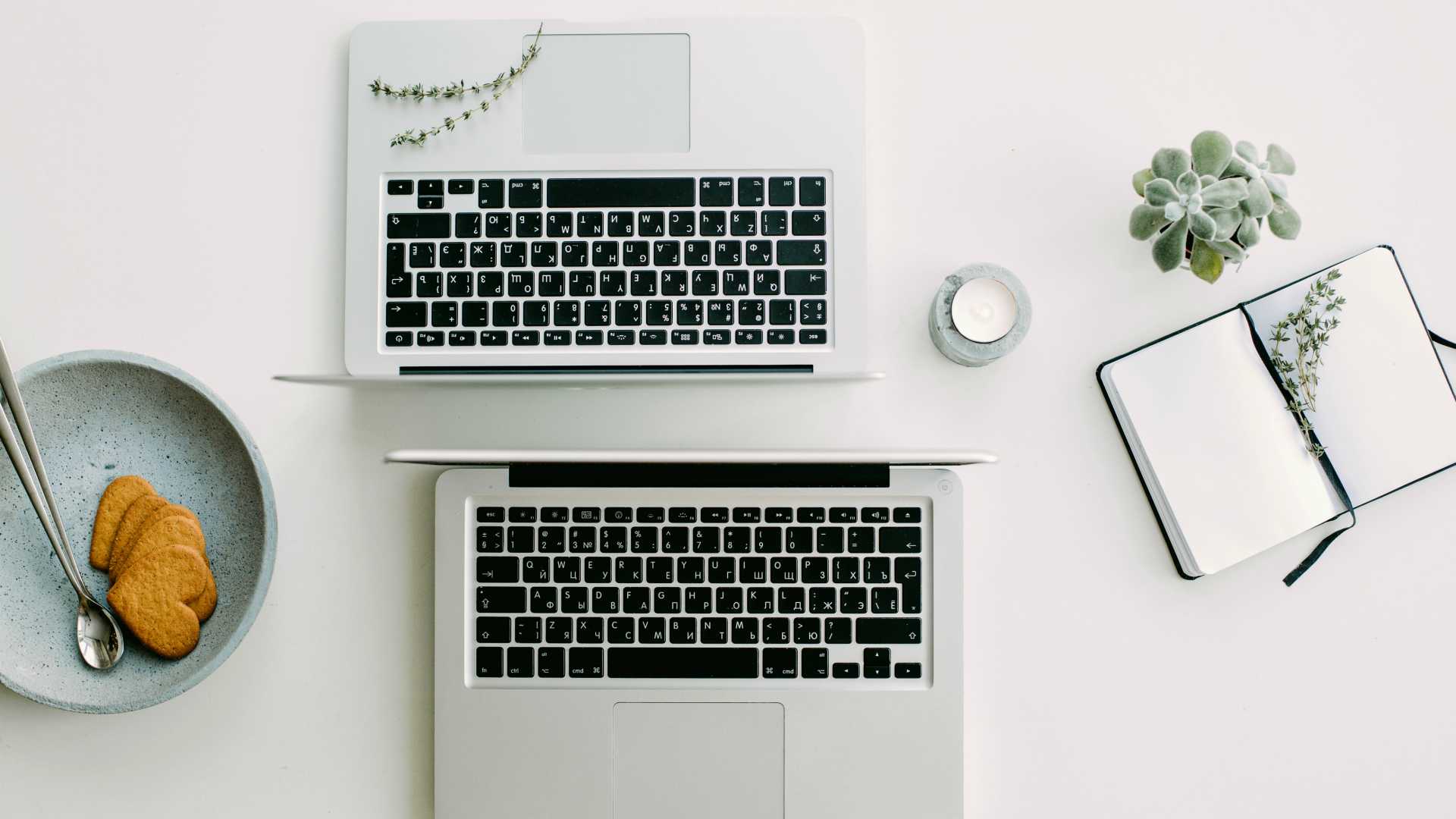 table with two laptops facing each other, a plant, an open notebook, a mug and a plate with two heart-shaped cookies on it
