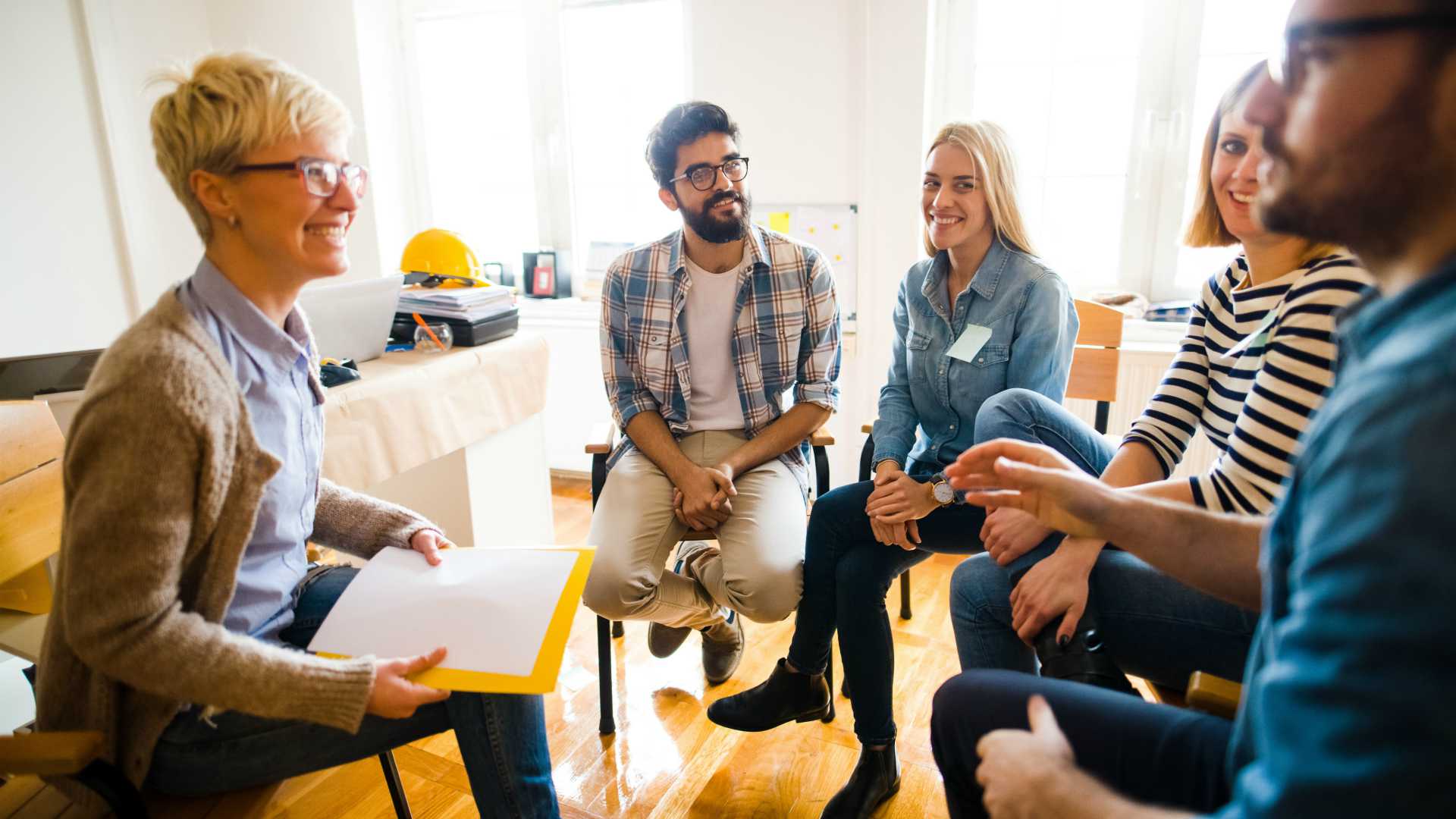 group of people sitting in chairs in a circle in an office; hard hat sitting on a desk in the background
