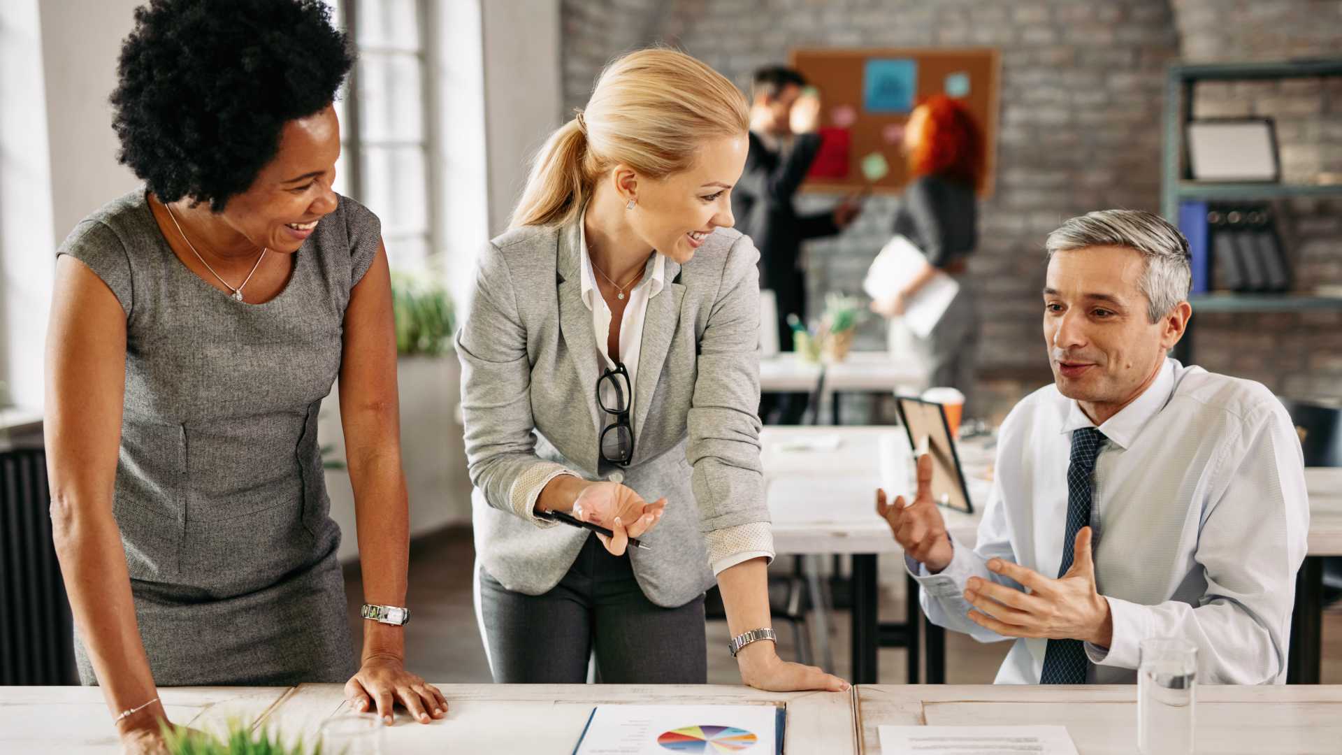 Group of smiling businesspeople communicating while working together in the office. There are people in the background.
