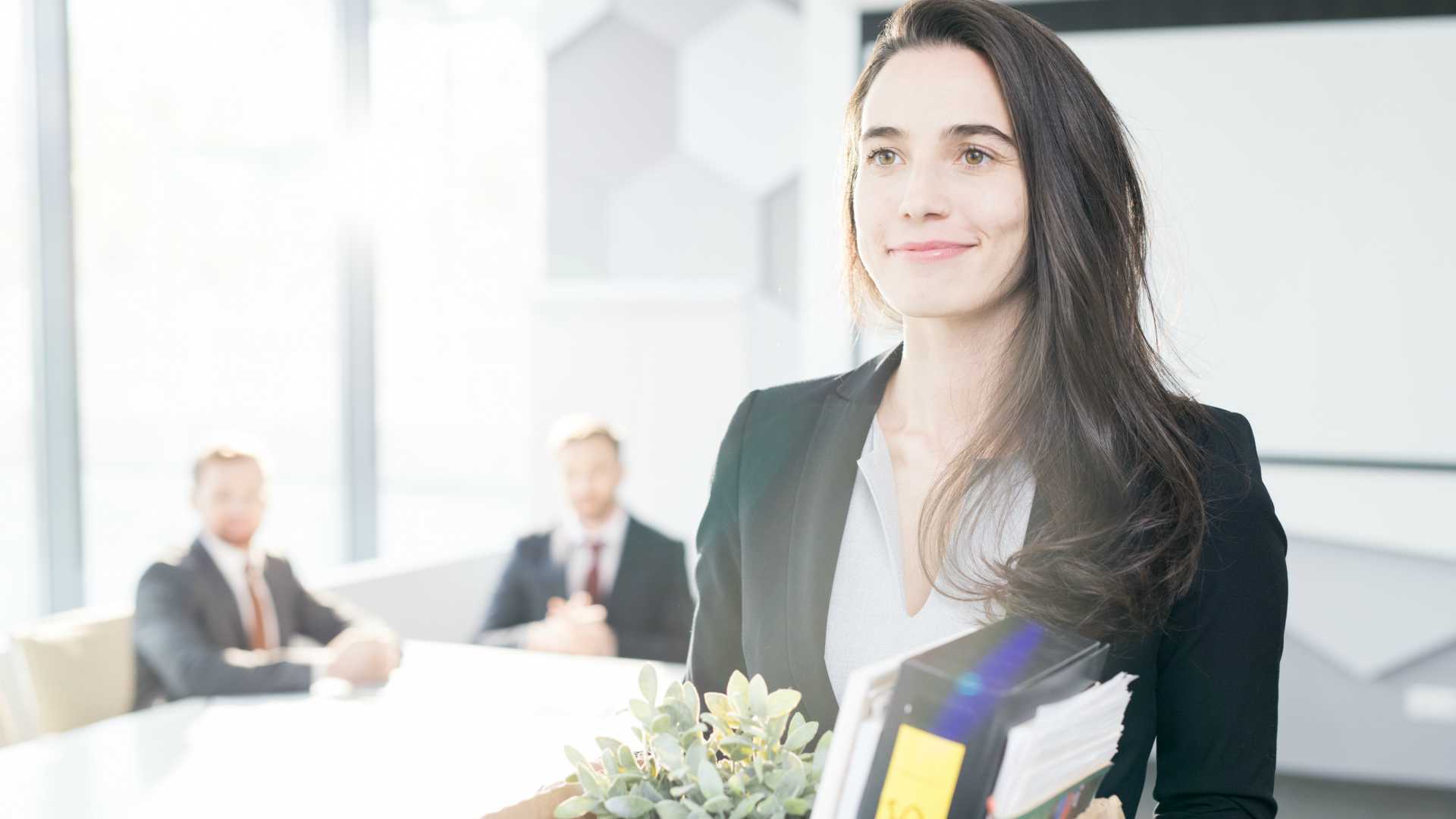 Young businesswoman leaving exit interview carrying a box of her things in brightly lit office