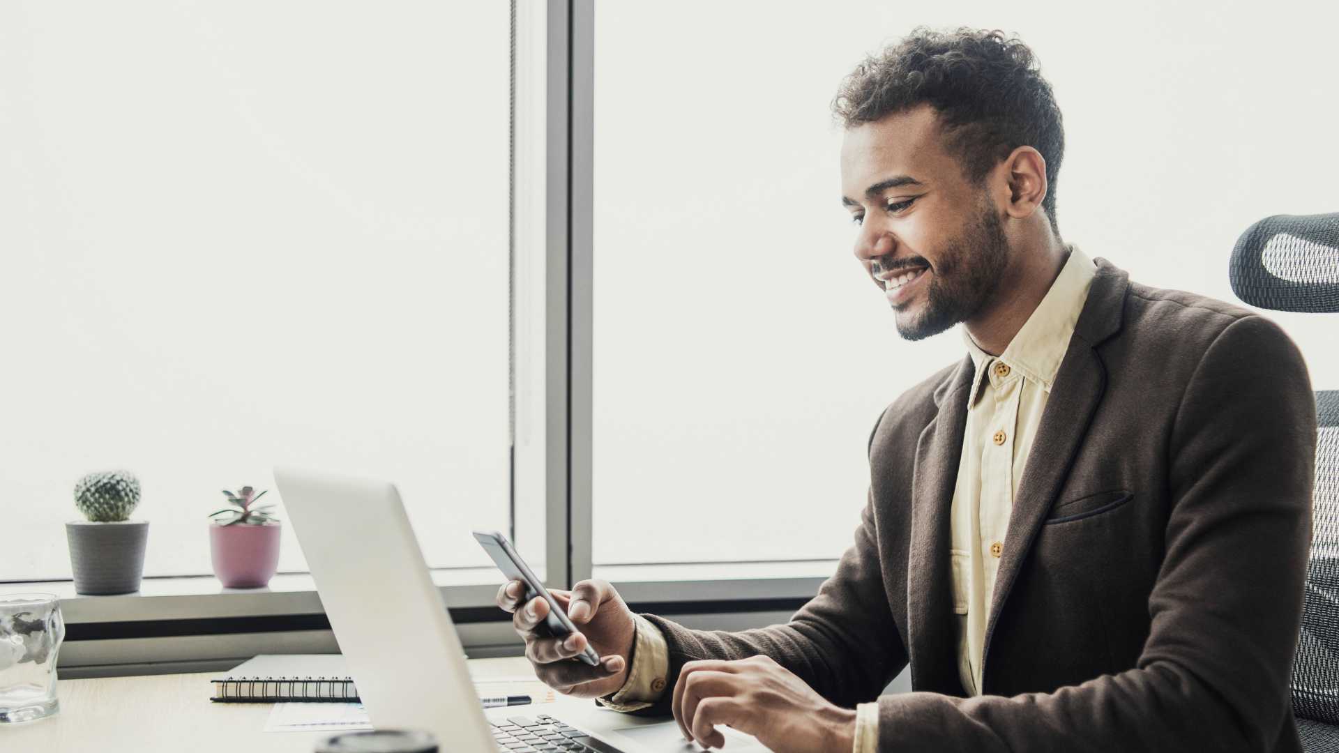 Businessman smiling at his phone while sitting at desk in front of laptop