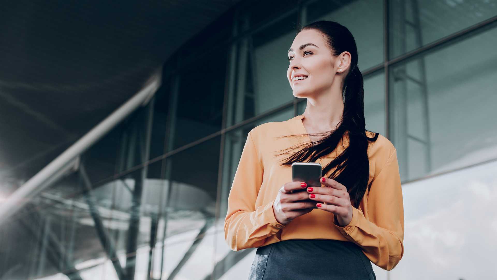 happy businesswoman holding a smartphone and looking to the distance, standing outside an office building