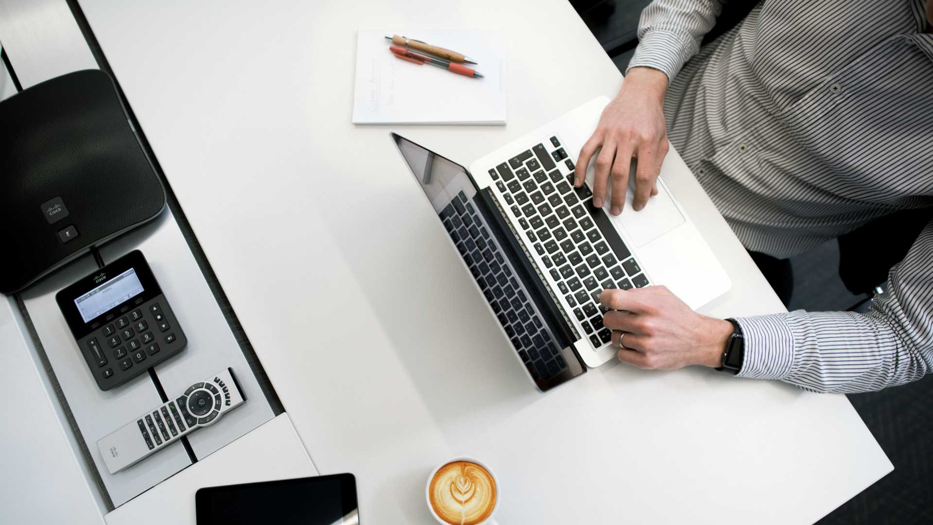 Aerial view of tidy desk with businessman sitting and working on laptop with a cappuccino close by