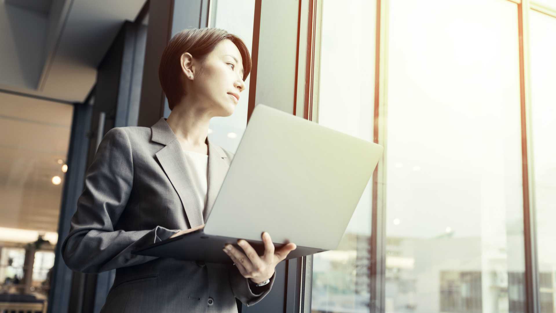 Young businesswoman thoughtfully gazing out office windoow and holding laptop