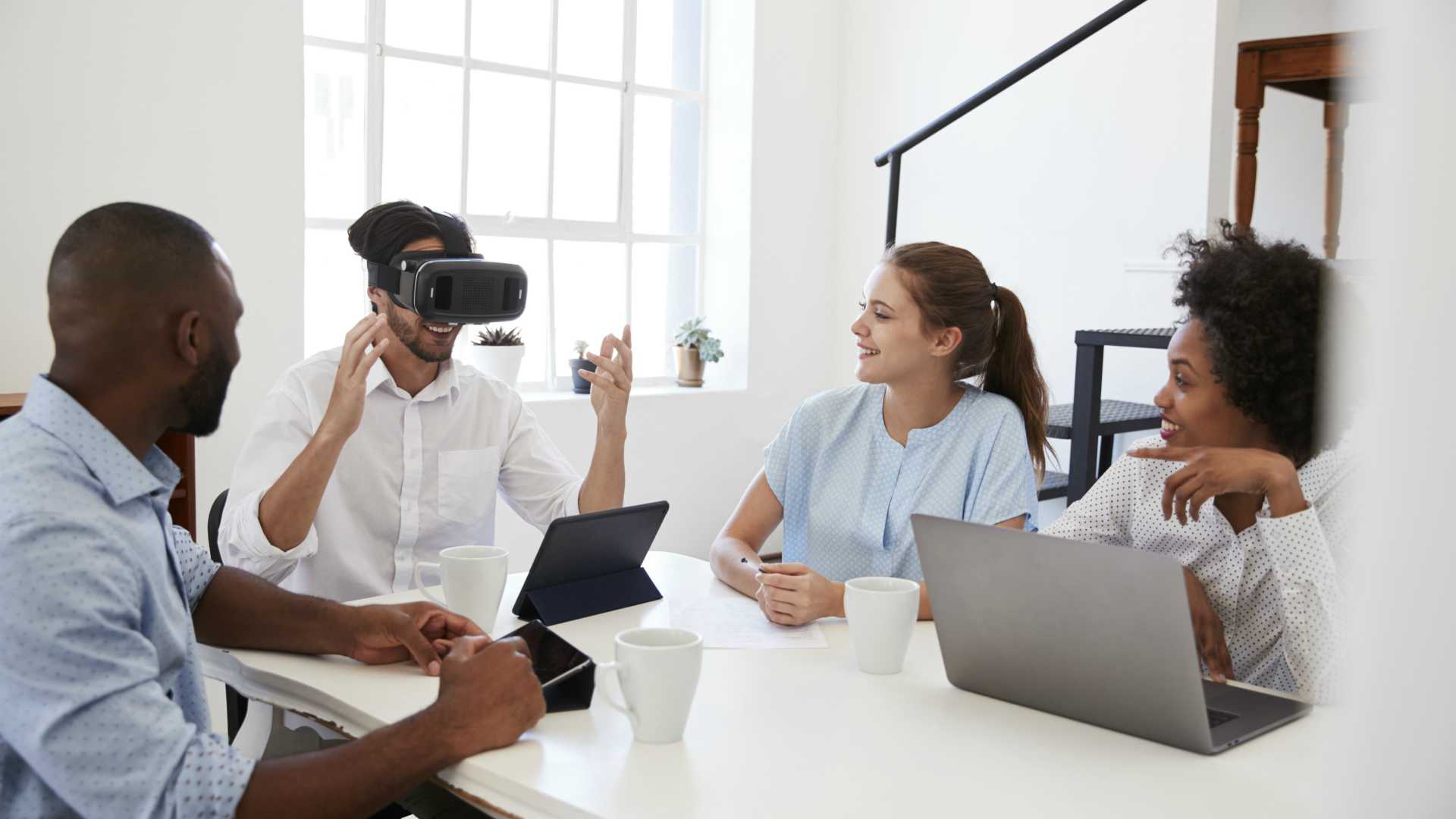 Man in VR goggles at a desk watched by colleagues in office