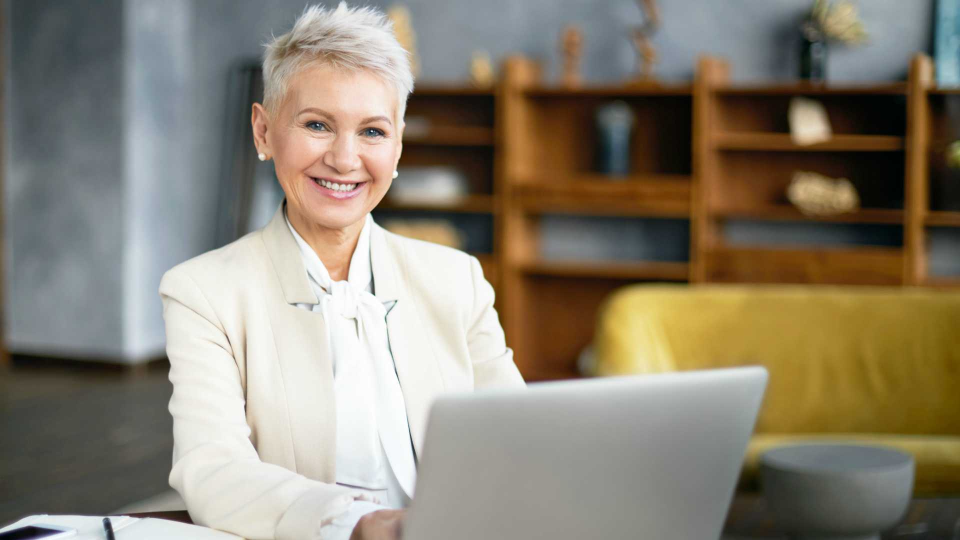 Grey-haired female executive working on computer and smiling at camera with home office in the background