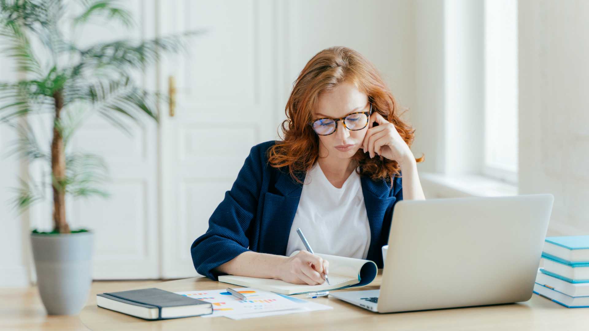 Red-headed businesswoman wearing glasses, working on laptop and jotting down notes at dining room table