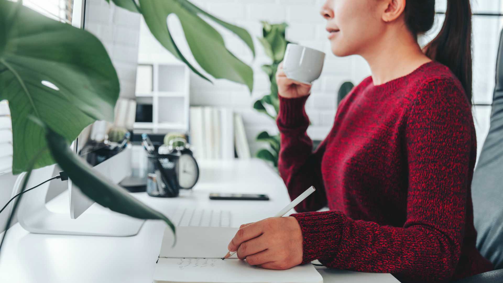 Businesswoman wearing red sweater sipping coffee from coffee mug and jotting down notes in notepad