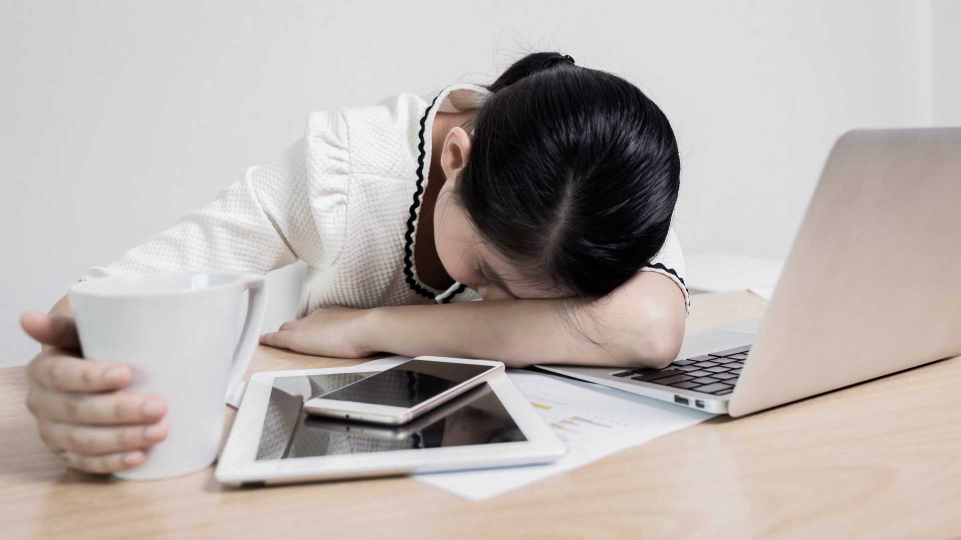 Business woman with head on desk in front of pile including tablet, smartphone and laptop, clearly stressed