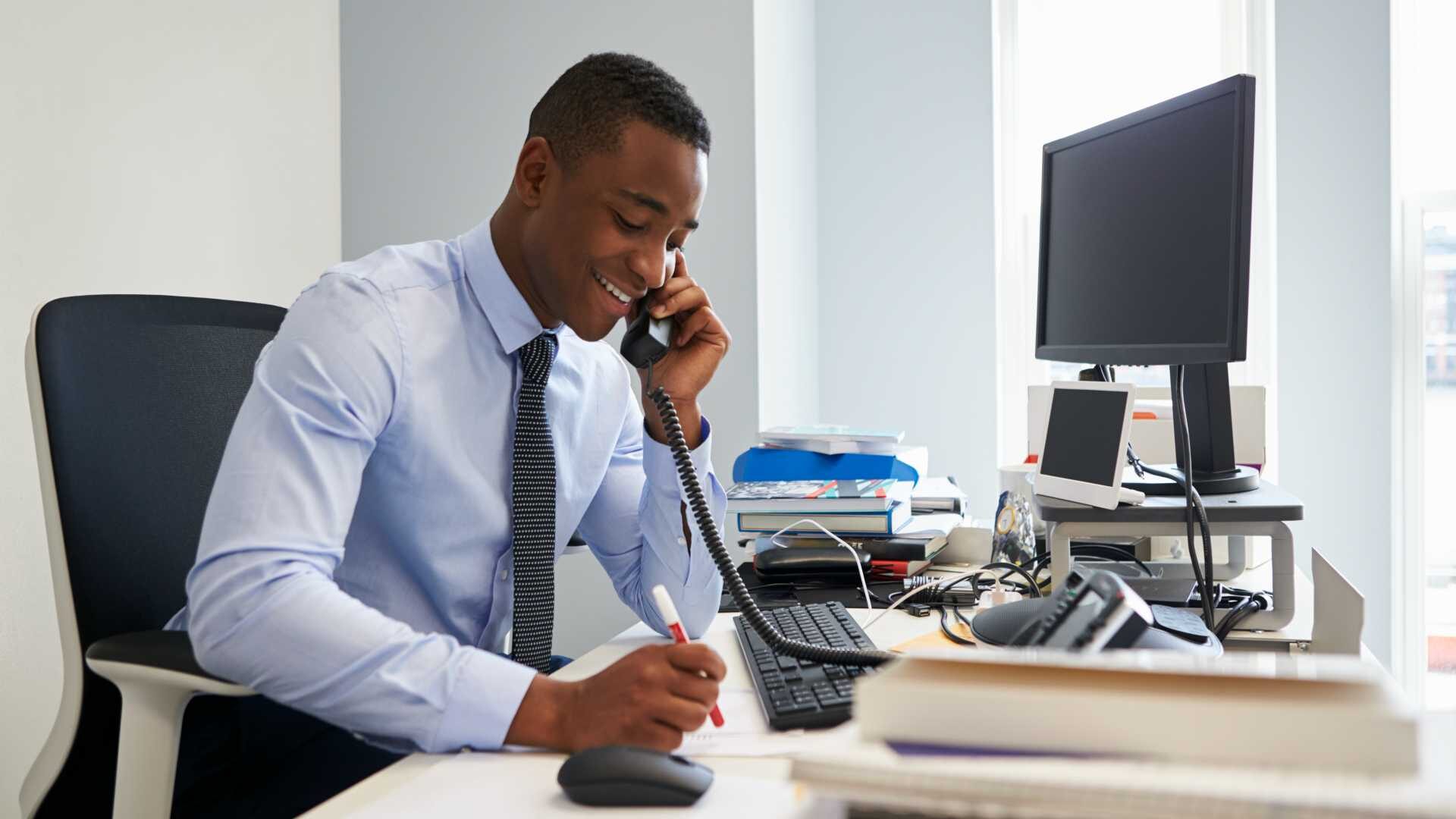A young Black businessman takes notes while on a call in his office