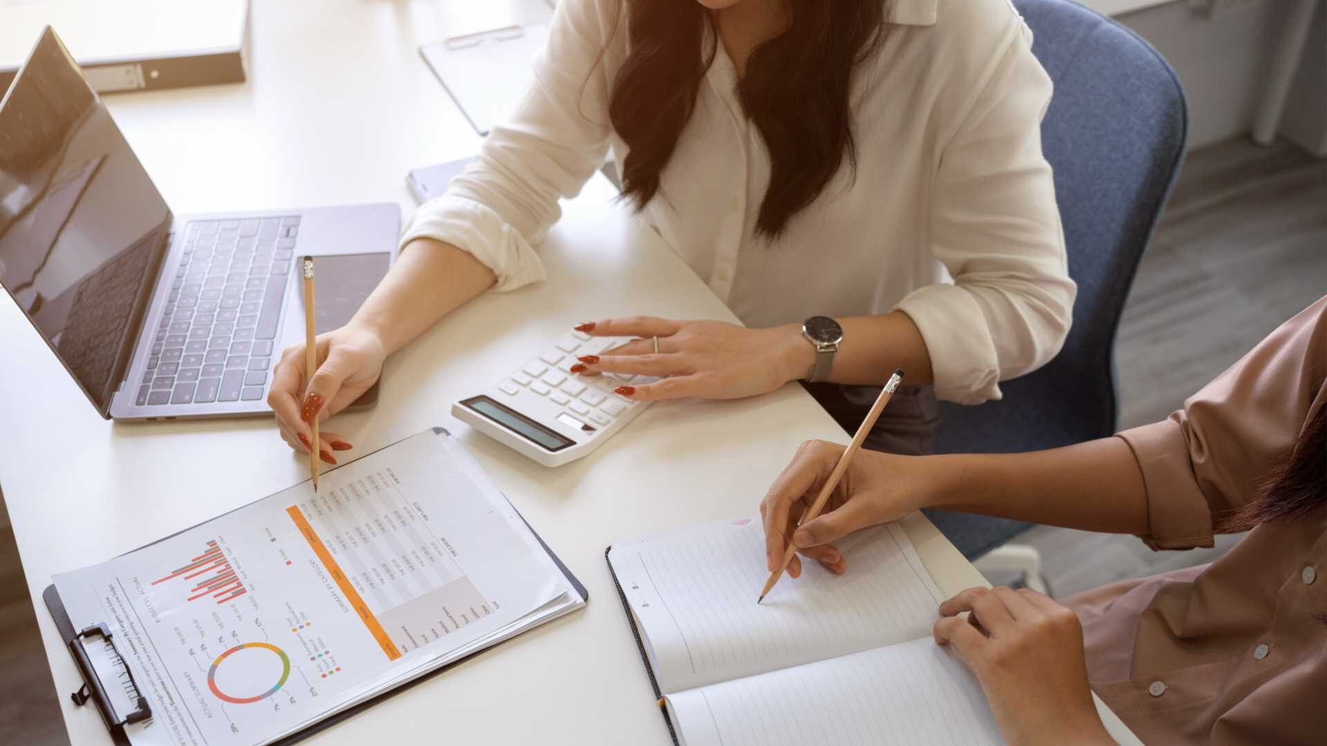 two women work at the same desk, one writing in a notebook and the other gesturing to a printout with her laptop open beside her