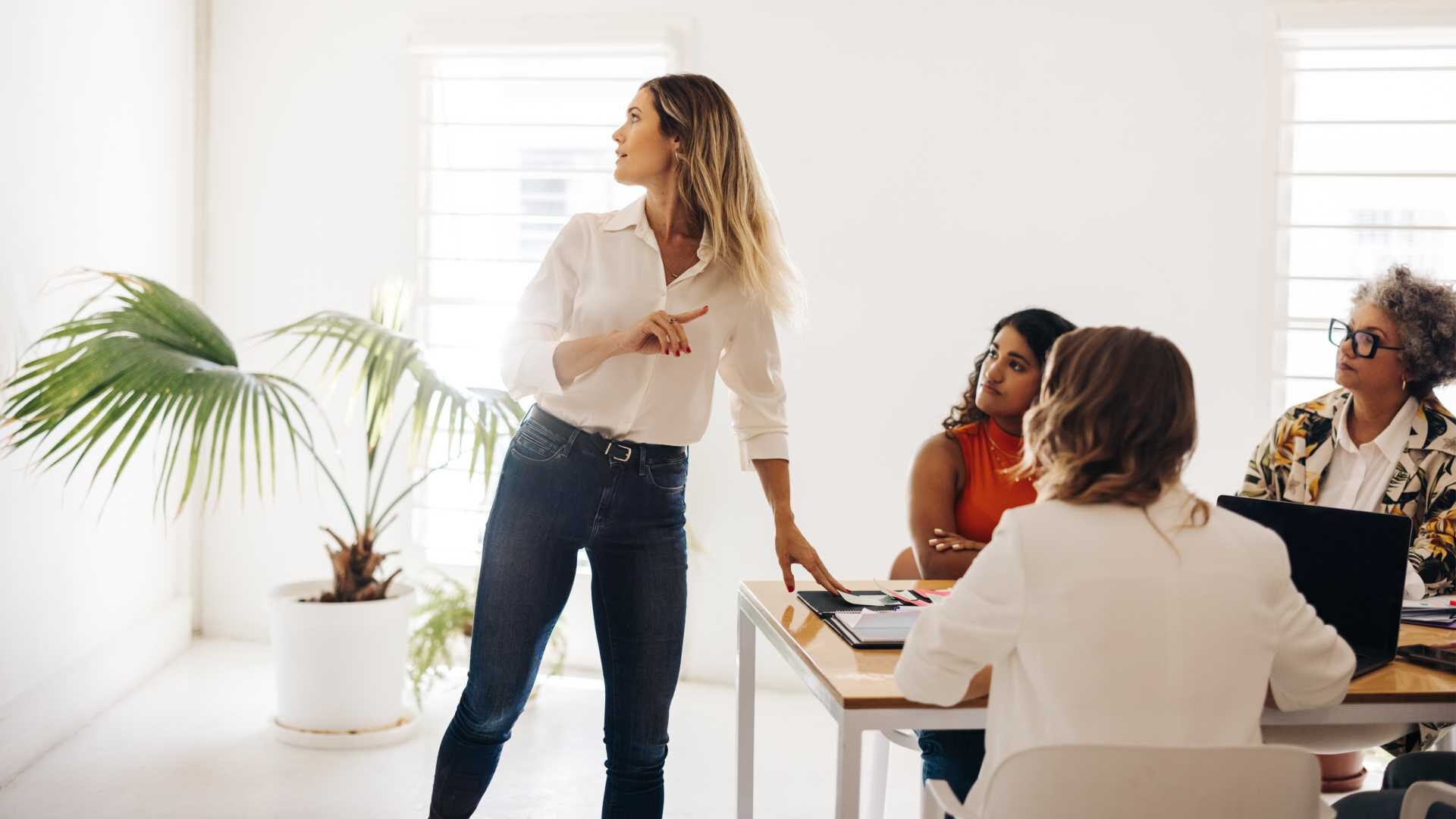 A standing business leader addresses her seated coworkers in a brightly lit workspace.