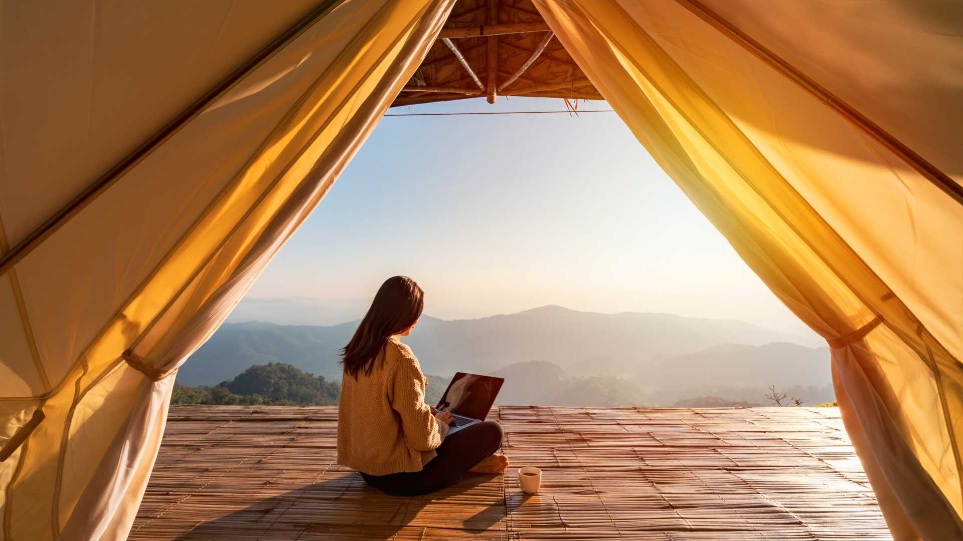A woman sits on the edge of a cliff looking out at the mountains next to a laptop.