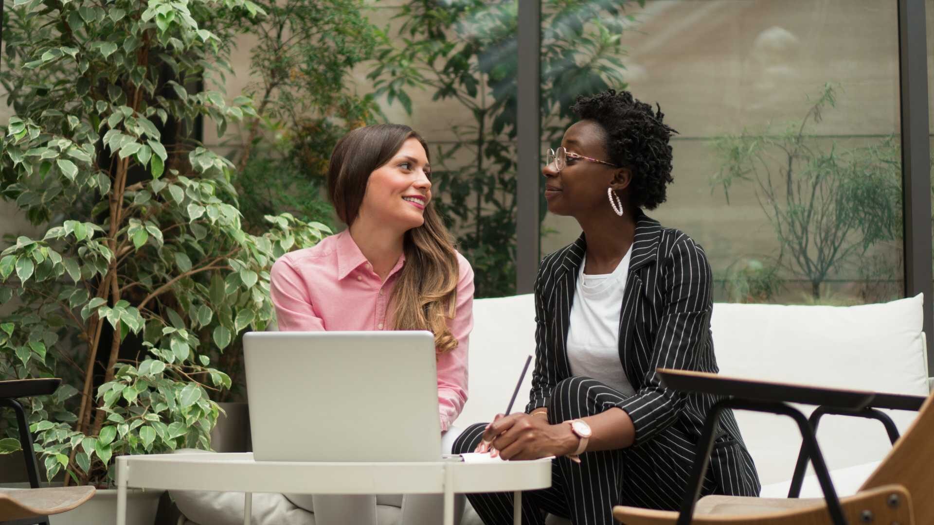 Two businesswomen discuss a project while looking at a laptop in an outdoor courtyard.