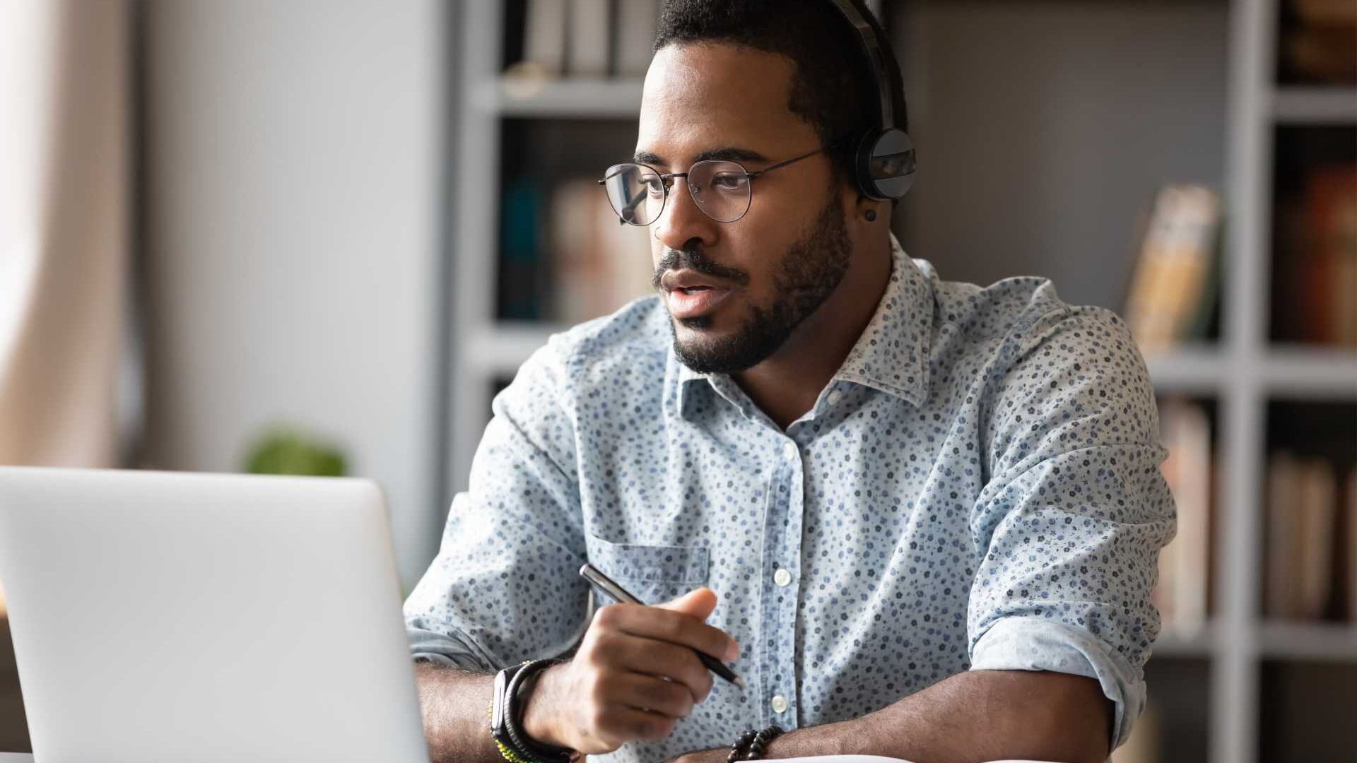 A man wearing a headset sits at a desk in front of a laptop while writing in a notebook in a home office.