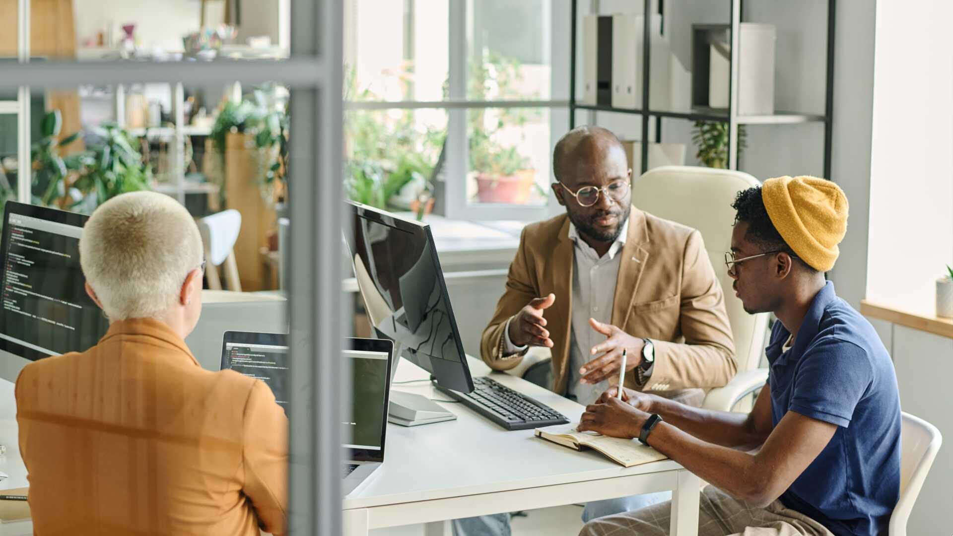 An experienced colleague speaks with a younger colleague, who is taking notes, in an office setting.