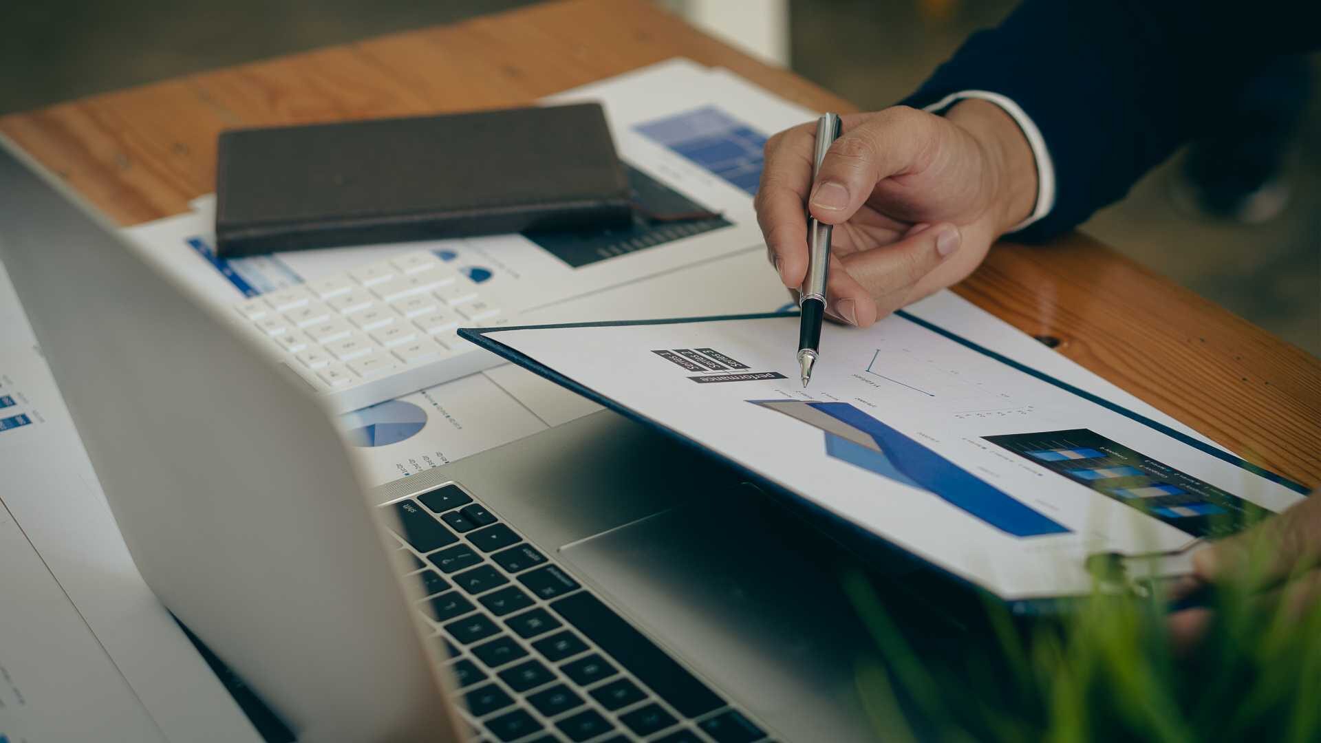 A businessperson holds a pen in front of an open laptop and a printed chart.