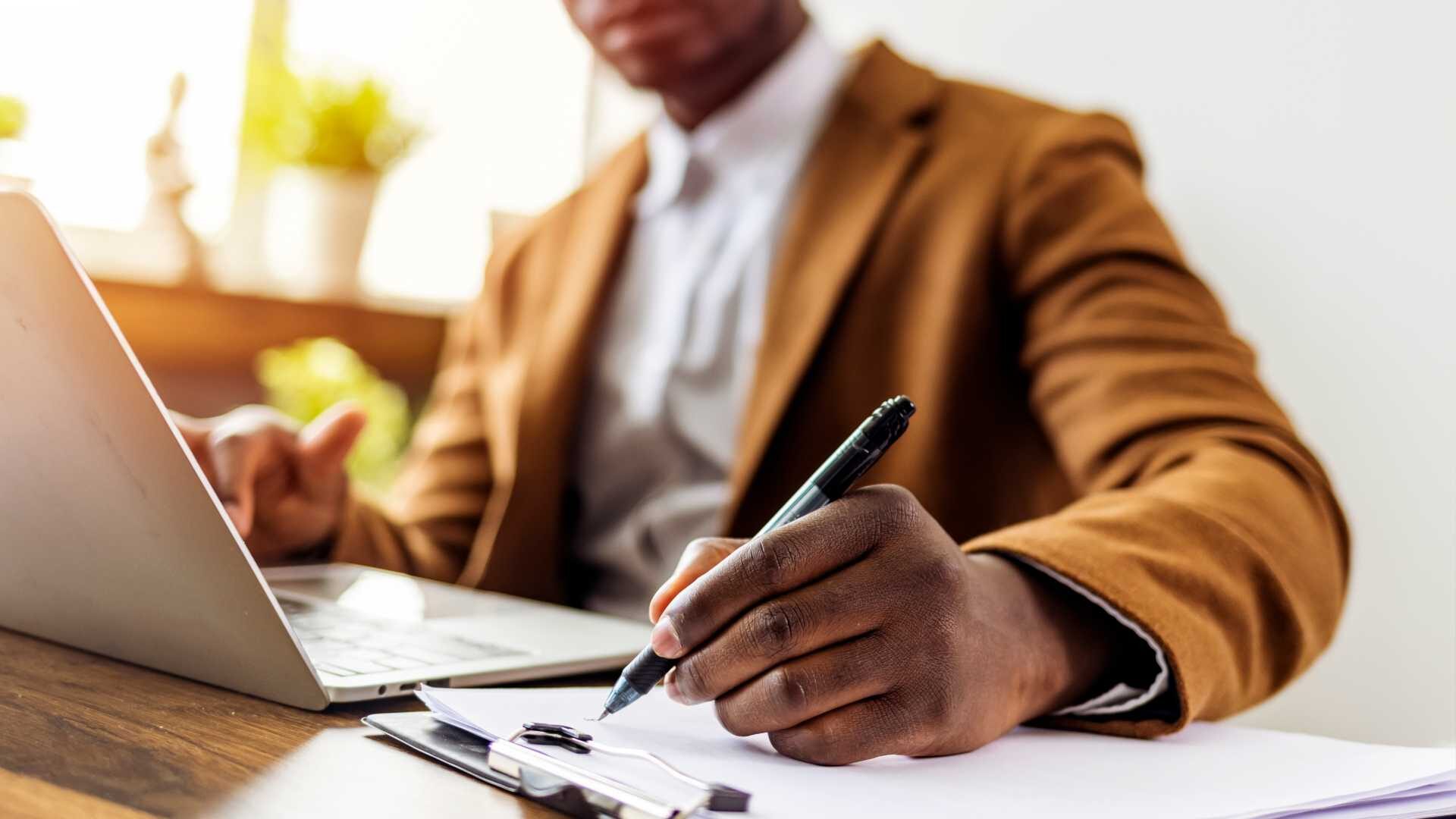 A working professional writes on a notepad while seated at a desk in front of an open laptop.