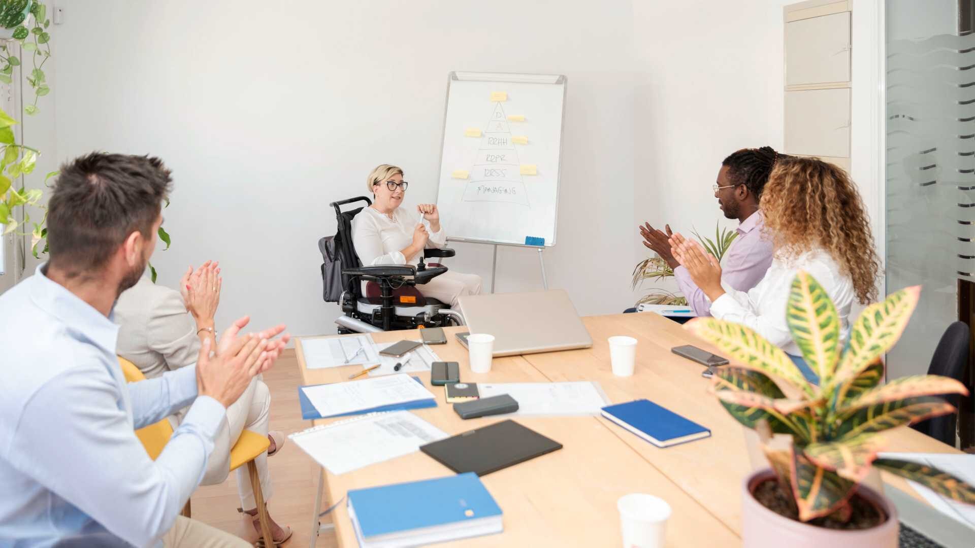 Woman with glasses in wheelchair presenting on a dry erase board to a table of clapping colleagues.