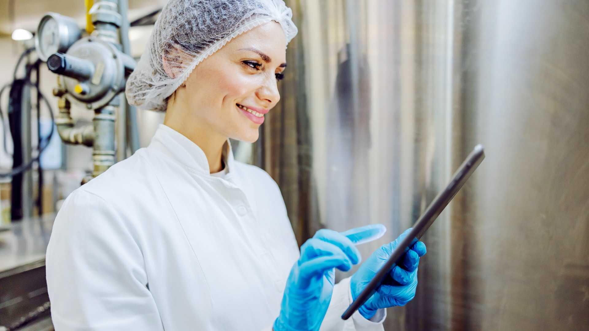 Woman wearing a hairnet, safety working attire and blue latex gloves at work holding a tablet.
