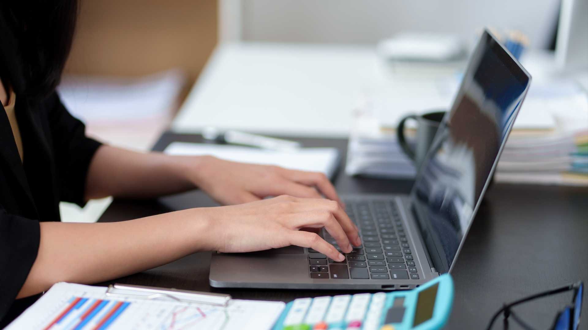 Closeup of woman's hands typing on a laptop. There is a calculator in the foreground, as well as some printouts.