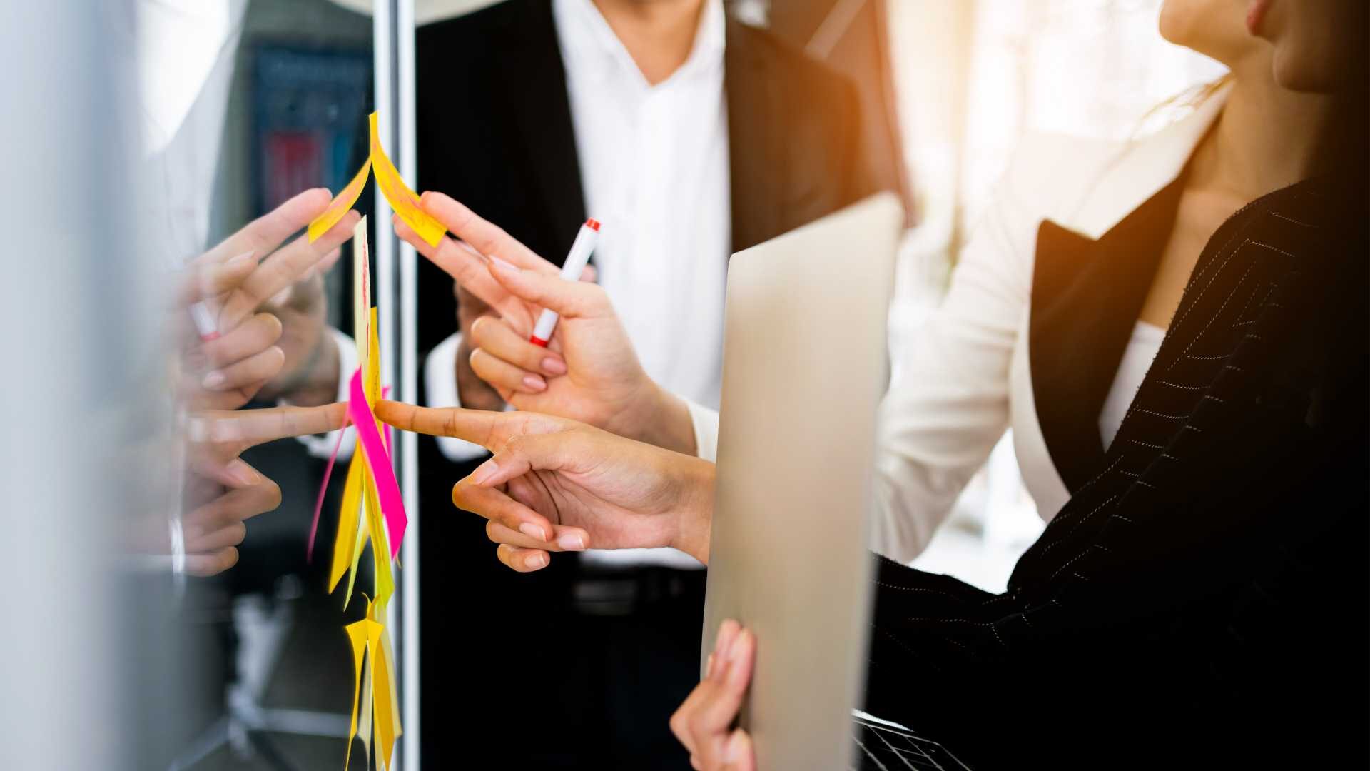 Business colleagues standing at a glass board with pink and yellow post it notes. Colleague holding a laptop.