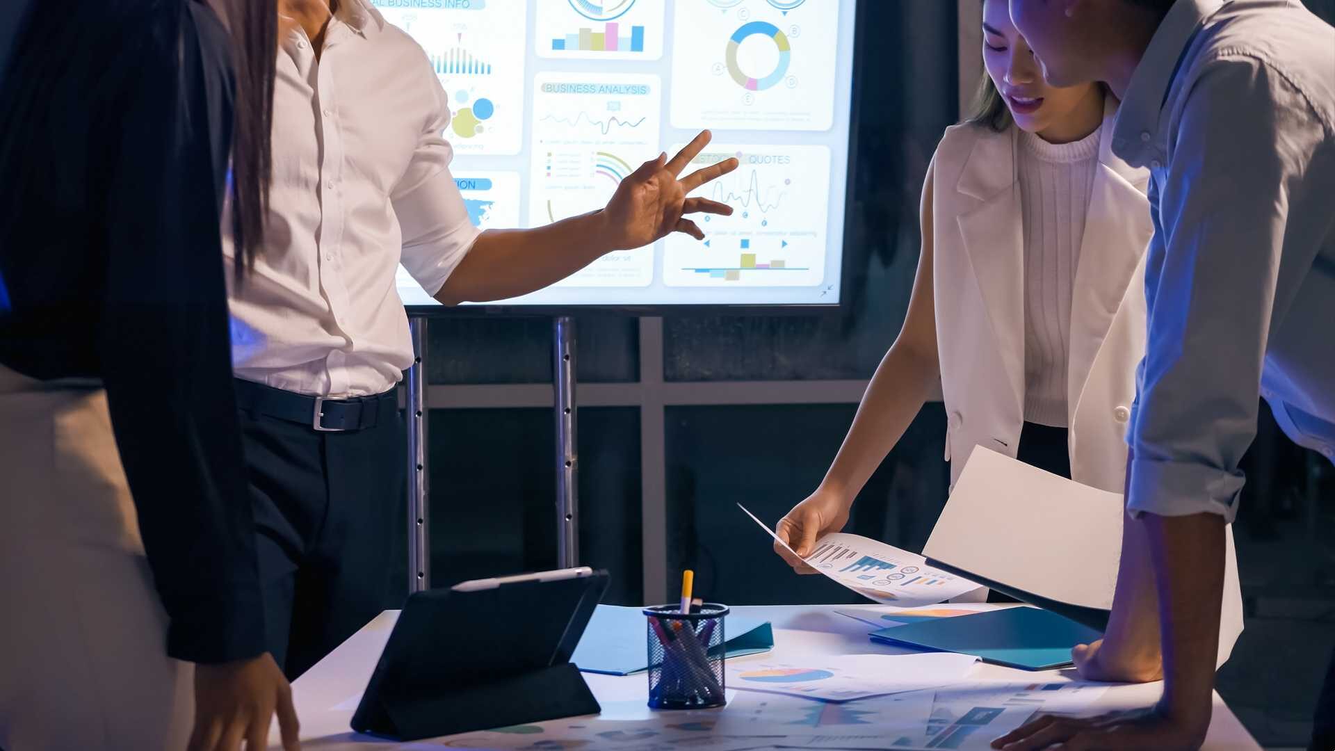 Colleagues standing around a table, measurements on a projector screen behind them, as they compare measurement documents and graphs.