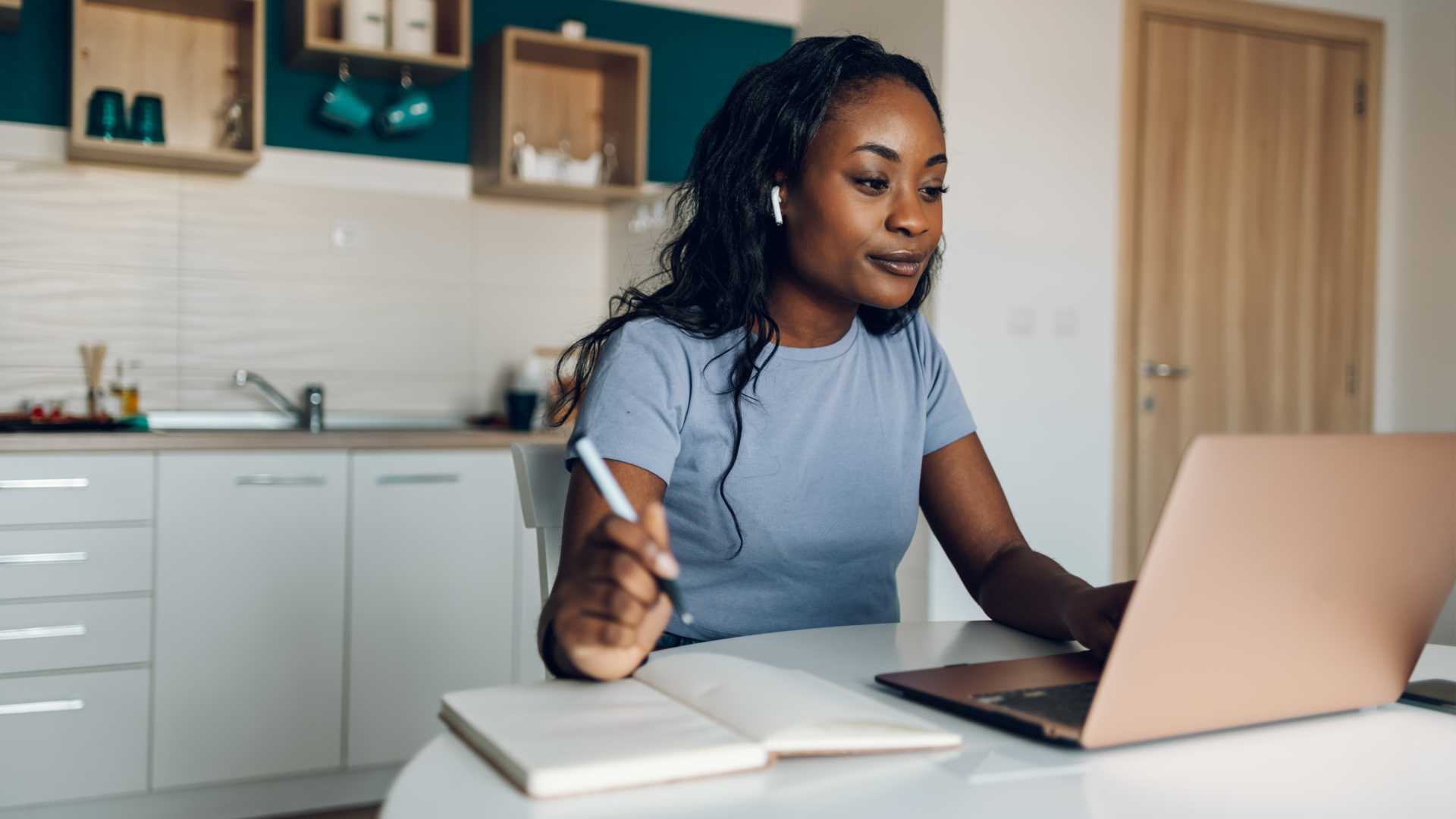 A young female professional works from home in front of an open laptop while taking notes in a notepad