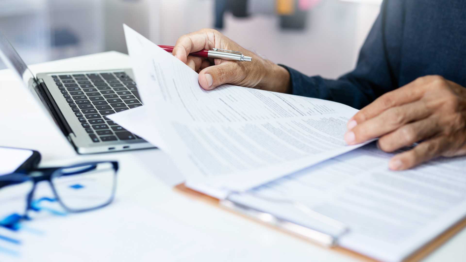 A businessperson reviews a printed resume while seated at a desk in front of an open laptop