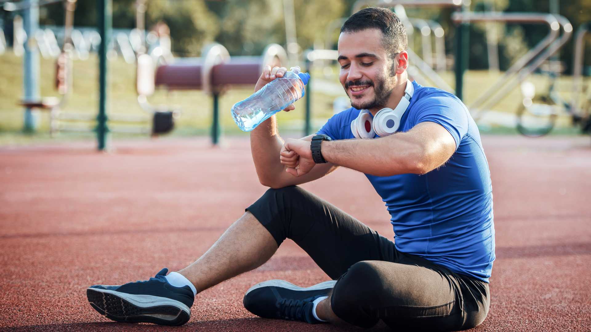 Track and field runner in workout gear and headphone around their neck, sitting on the track with water bottle.