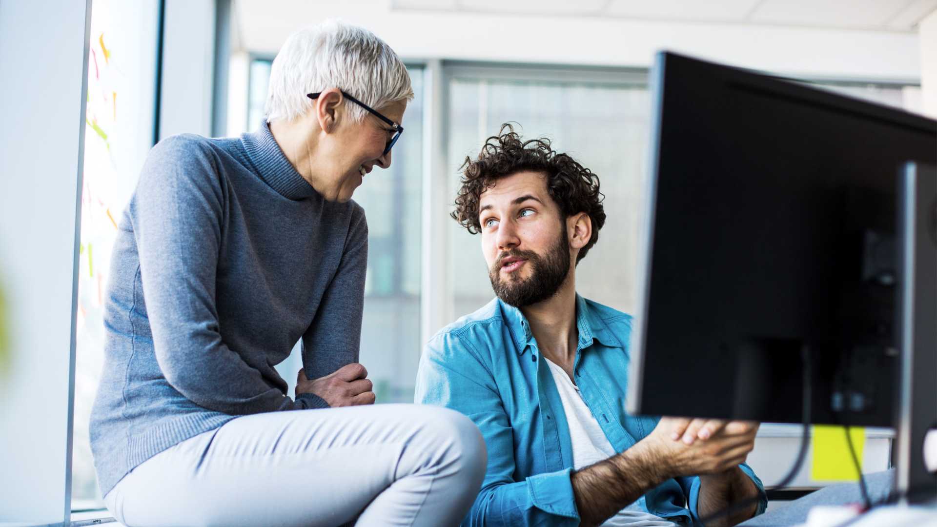 Two colleagues engage in a conversation in an office setting in front of a computer.