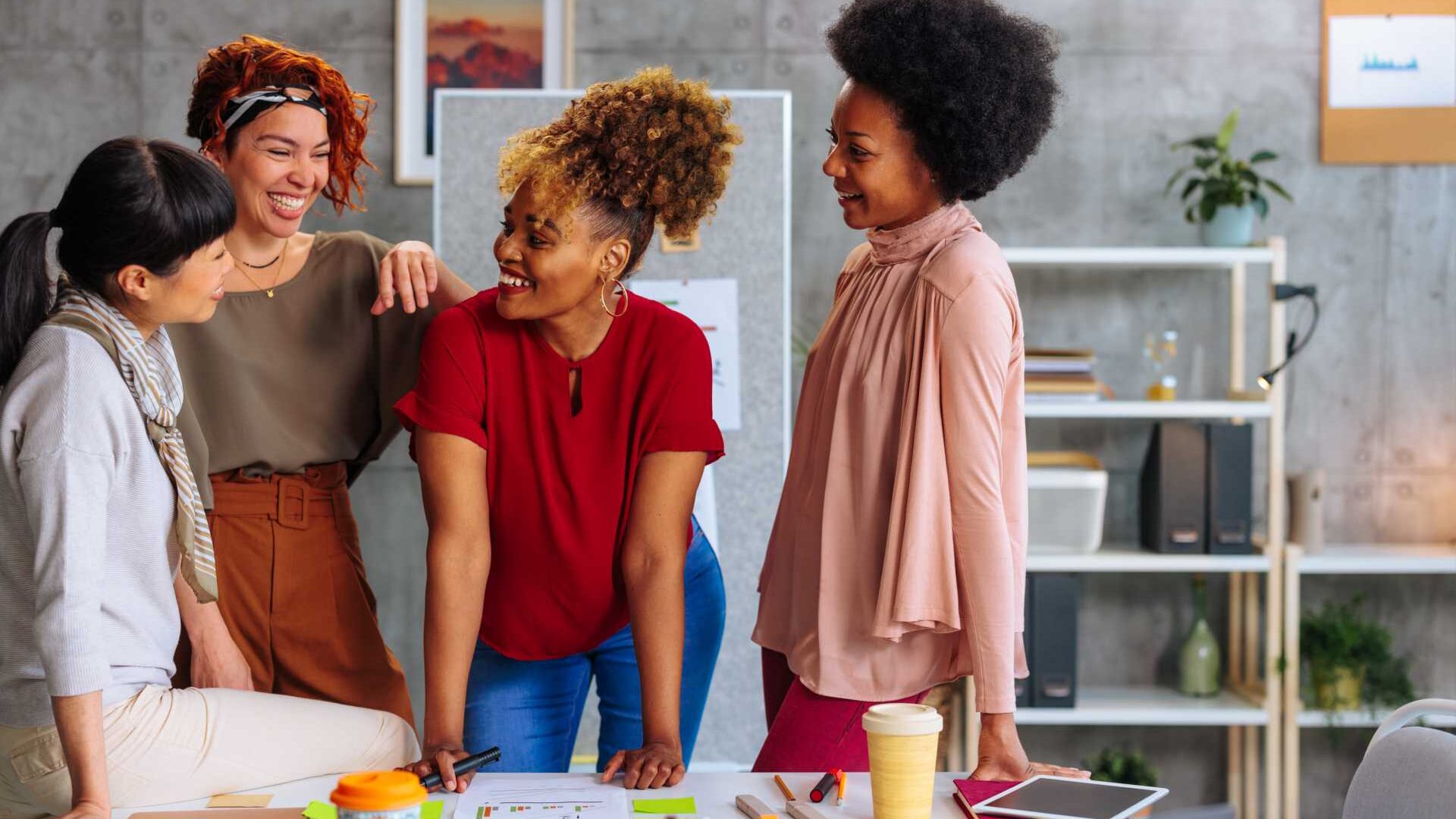 A group of women collaborate in a workplace setting in front of a desk with coffee cups, pens and pencils