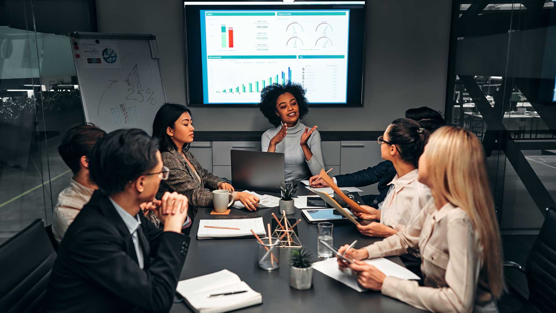 A small business meeting takes place around a conference room table with a TV displaying charts and graphs
