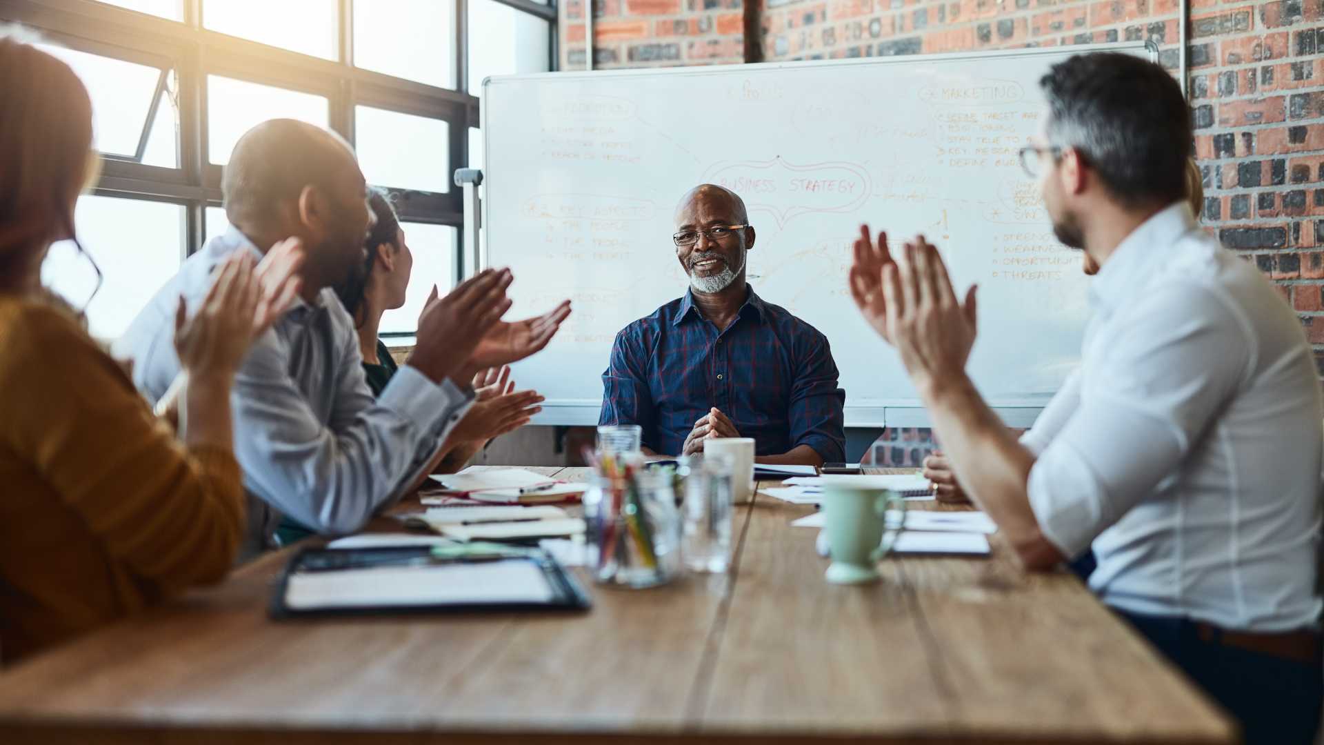 A businessman sits at the front of a conference room table surrounded by colleagues clapping