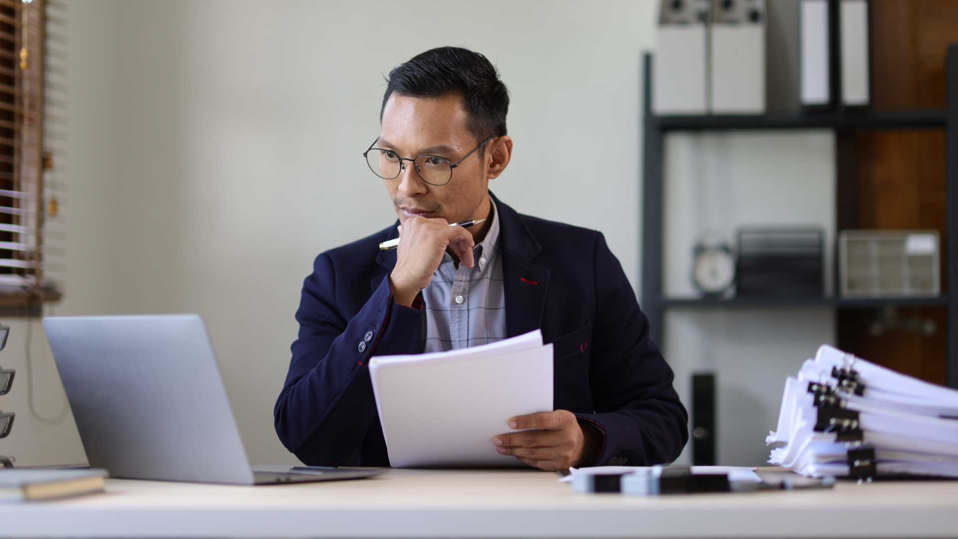 Businessman seated at an office desk holding a piece of paper looking pensive.
