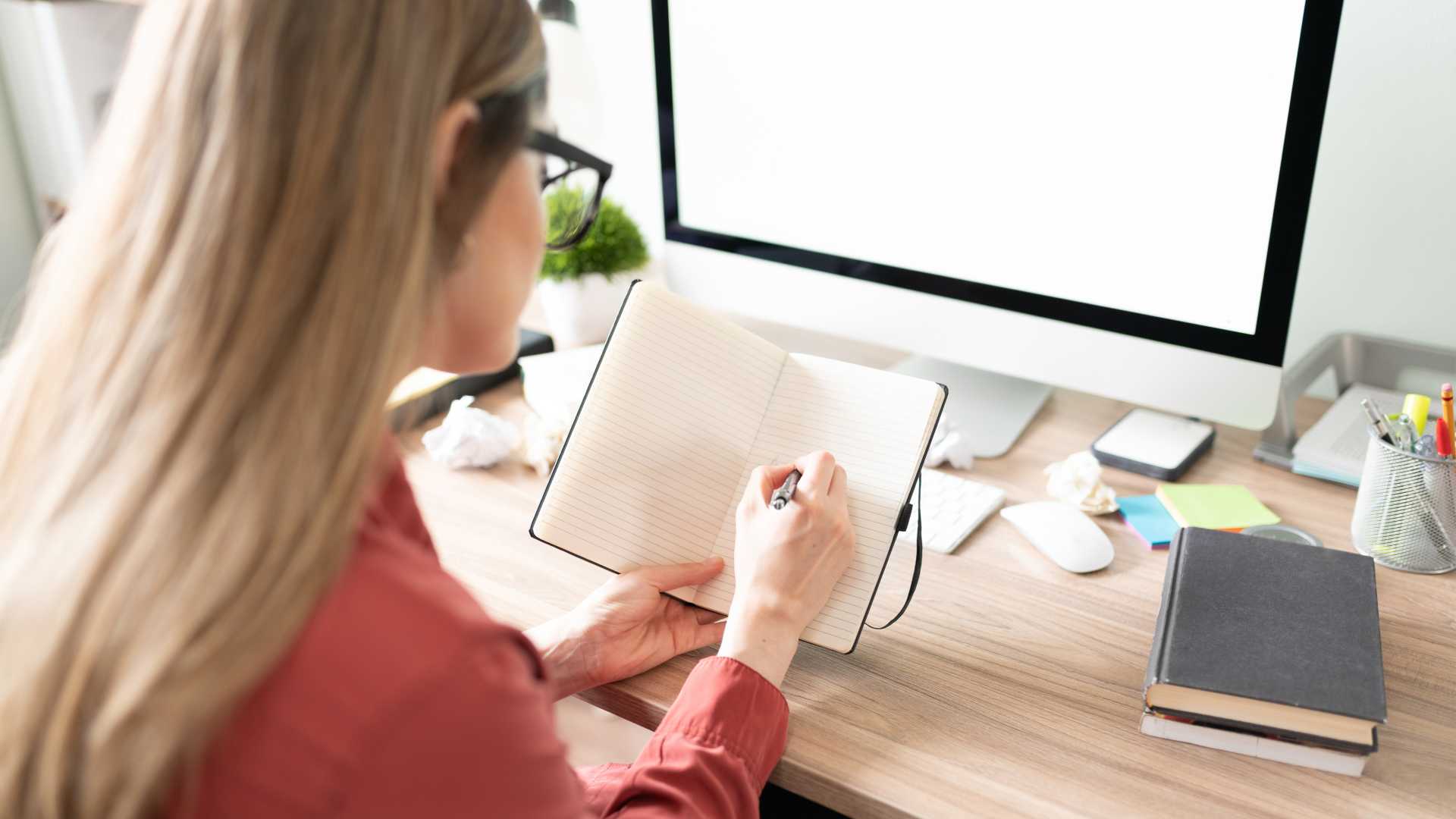 A woman seated at a desk in front of an open computer writing in an open notebook.