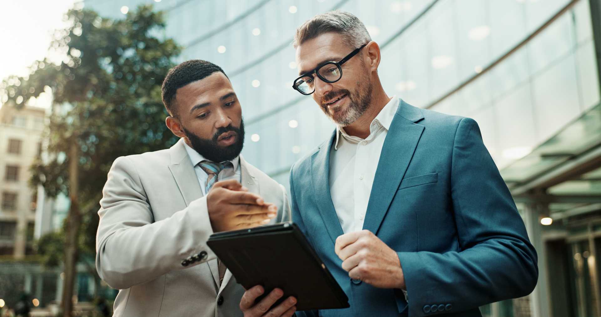 Two men in business attire standing outside a building looking at a tablet