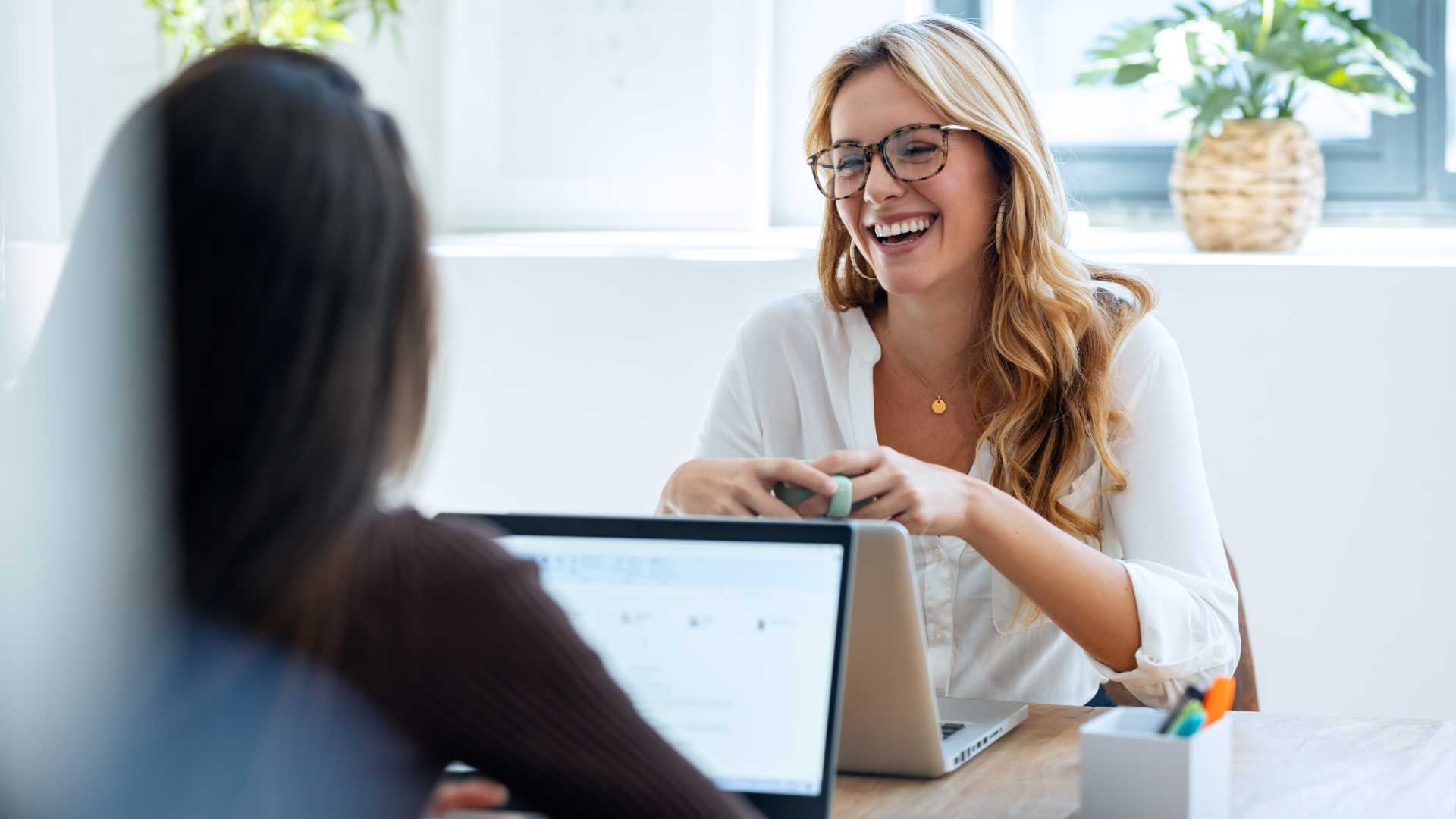 Two businesswomen having a conversation in a work setting in front of open laptops