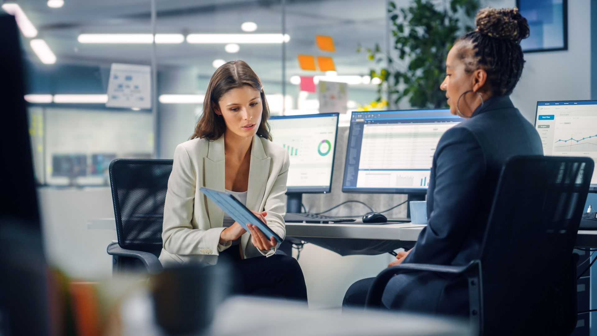 Two professionals sit in a modern office discussing information on a tablet, with computer monitors displaying charts and data in the background.
